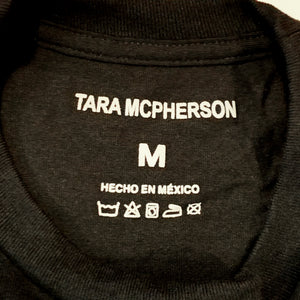 Close-up of the inside collar of a black crewneck sweatshirt, showing white printed text that reads 'TARA MCPHERSON', size 'M', 'HECHO EN MÉXICO', and care symbols.